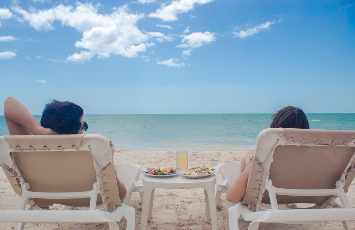 Two people relax on sunbeds with refreshments by the sea, enjoying a sunny summer day.