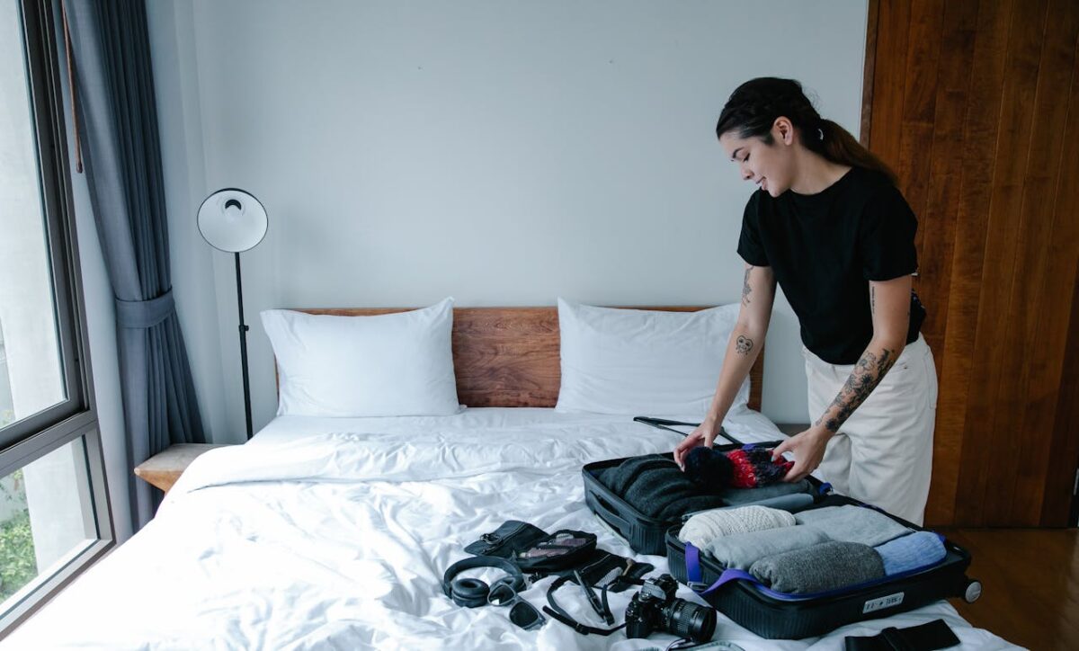 Woman packing suitcase with clothes and camera in a bright bedroom.
